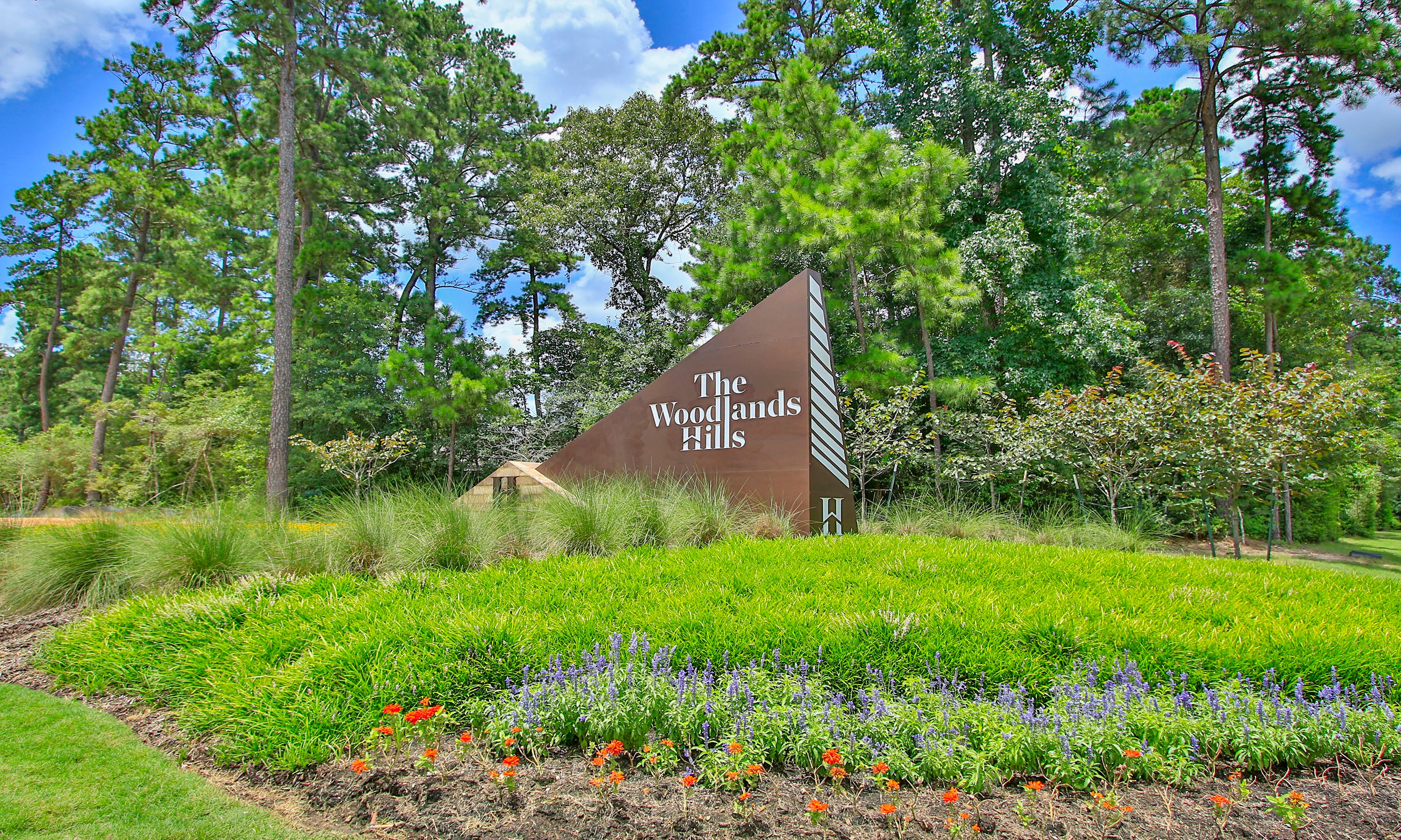 "The Woodlands Hills Entrance Sign Amidst Nature's Beauty" "The Woodlands Hills entrance sign surrounded by lush greenery and vibrant flowers in a wooded area."