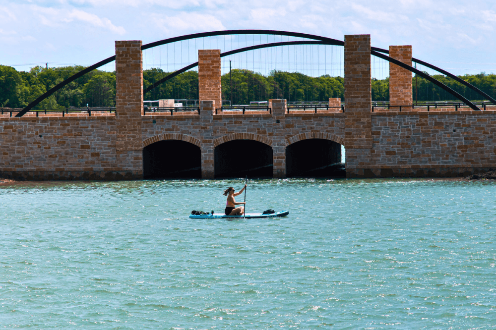 Elegant Stone Bridge Over Lake in Solterra Texas Community Stone bridge with arched supports over lake, paddleboarder in foreground, lush greenery, Solterra Texas community.