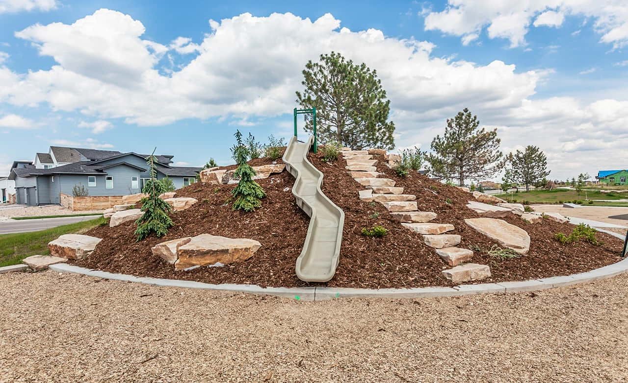 Trailside on Harmony Playground with Slide and Landscaped Hill Modern playground with slide on landscaped hill, Trailside on Harmony, featuring boulders and lush greenery under blue skies.